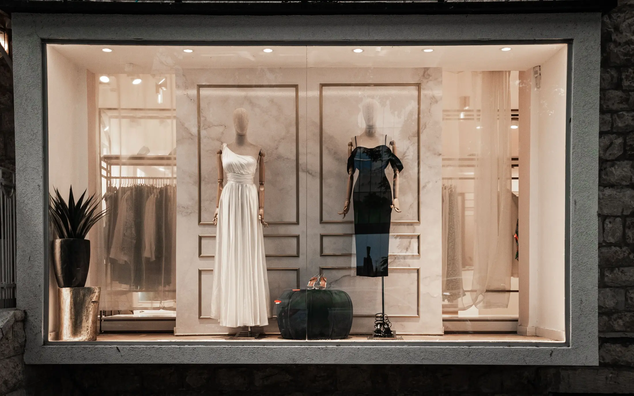 Boutique storefront window with mannequins in a white gown and black dress at dusk