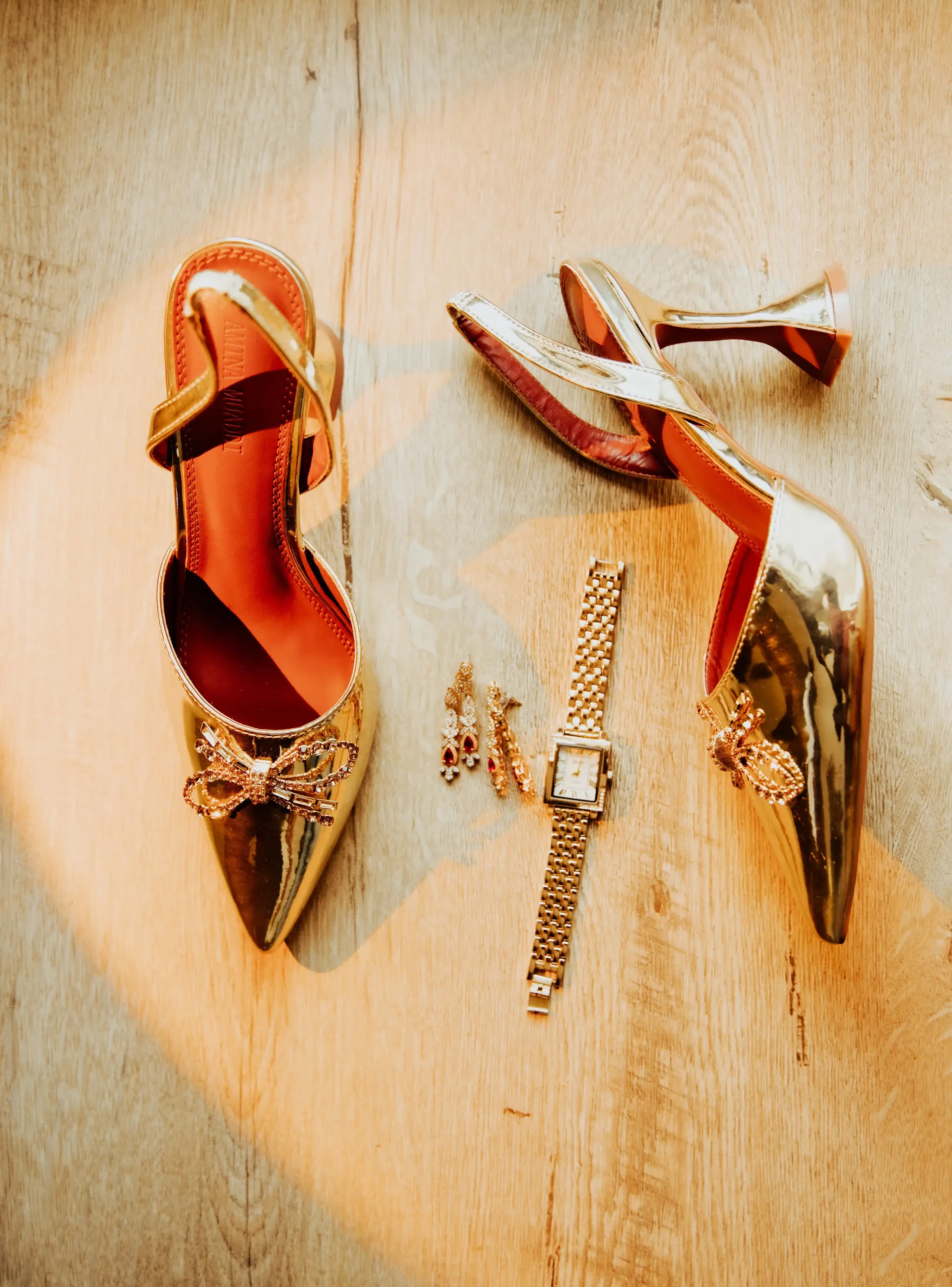 Gold heels, a wristwatch, and earrings arranged on a sunlit wooden floor