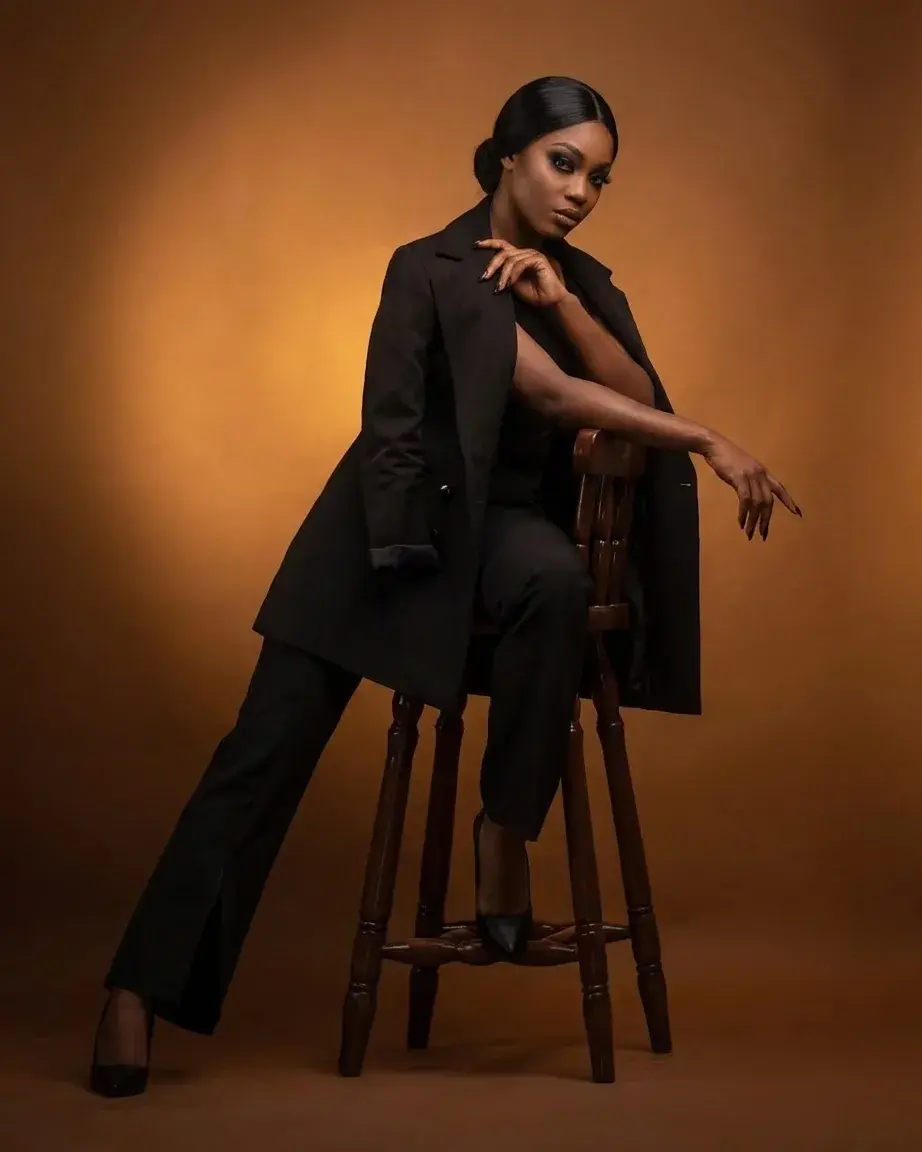 A woman in a black draped coat seated on a stool against a warm amber studio backdrop