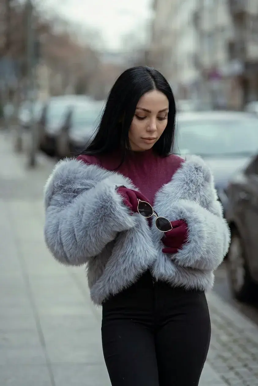 A woman in a grey faux fur jacket and burgundy turtleneck on an overcast city sidewalk
